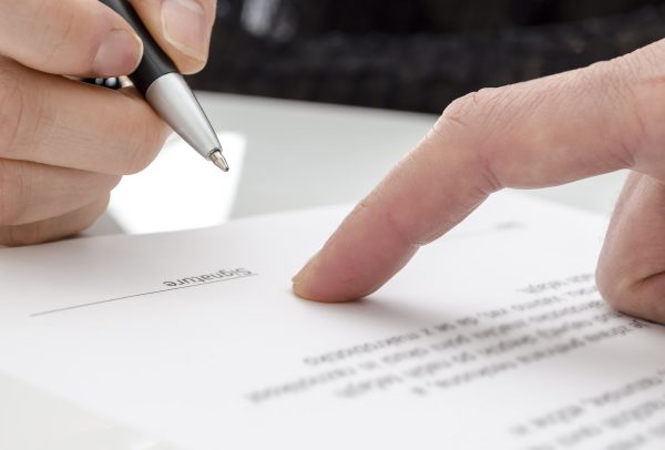 Detail of a woman signing a paper. Male finger showing where to sign.